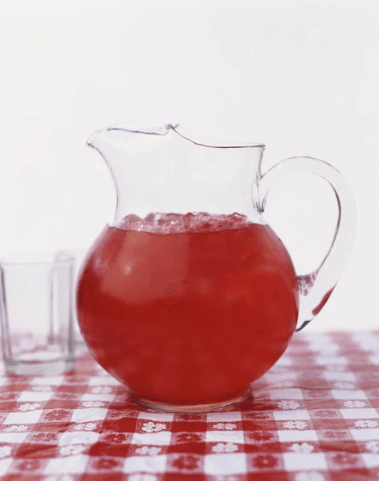 A glass pitcher with a red liquid and ice cubes sits on a red-checked tablecloth.