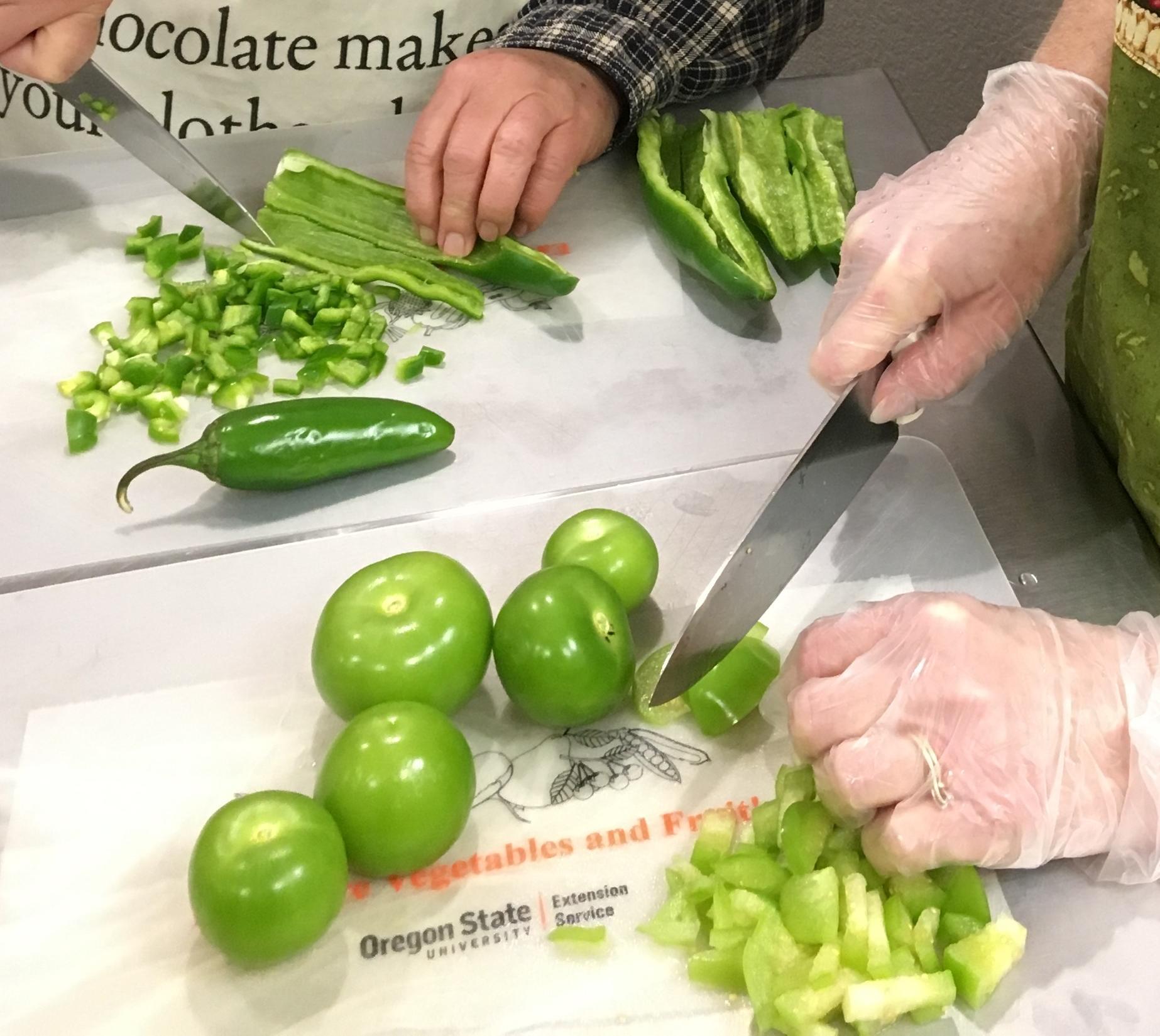 Close-up of the hands of two people, both with knives; one cutting peppers, the other cutting tomatillos.