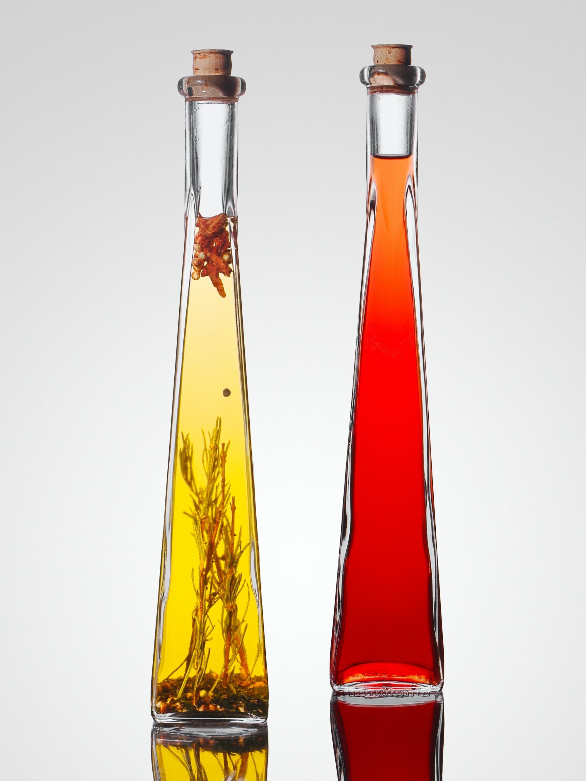 Two long, slender bottles containing liquids, one yellow with herbs visible inside; the other red with no additions.