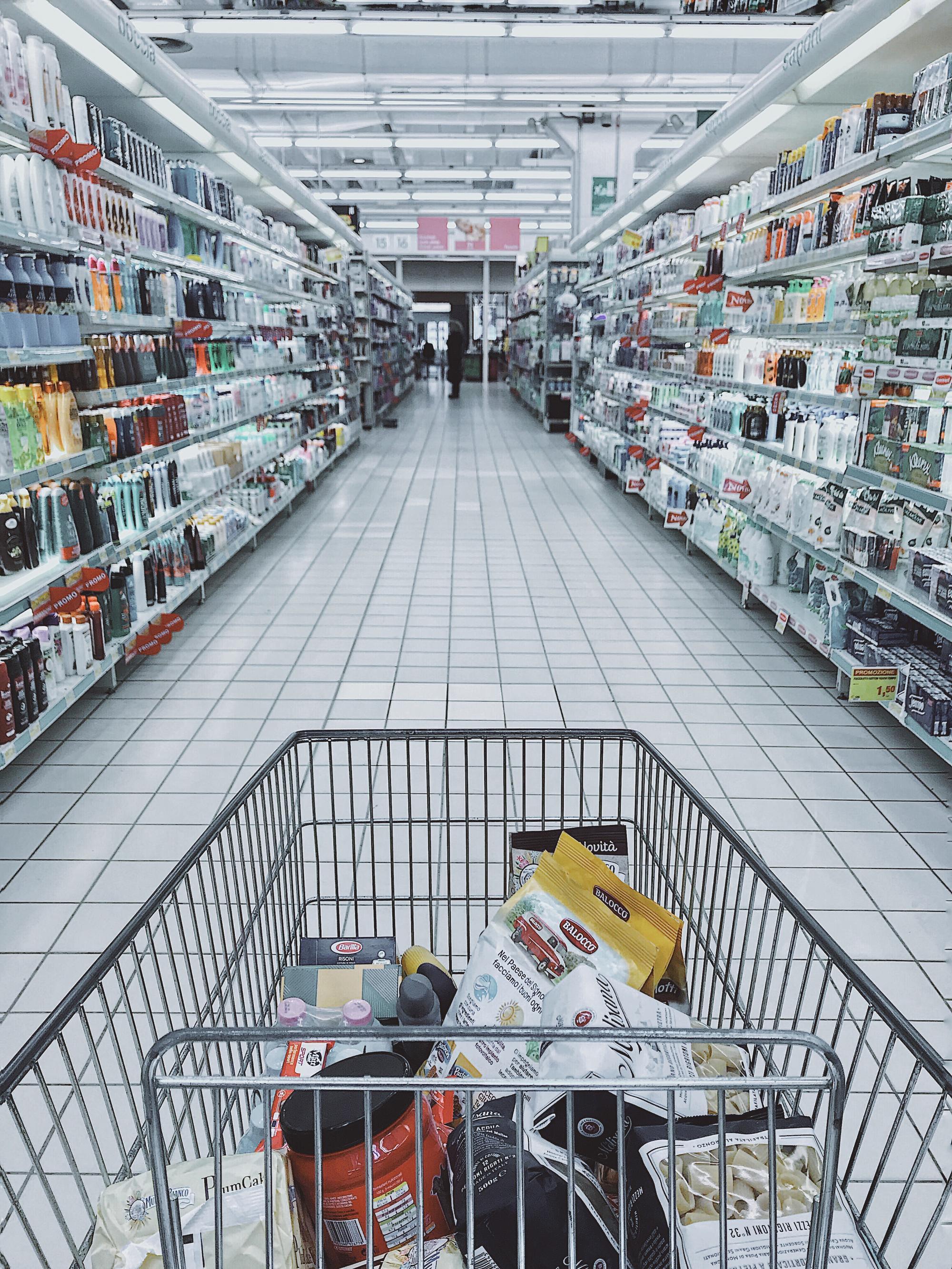 A partially filled shopping cart is depicted in the middle of an aisle between shelves in a store.