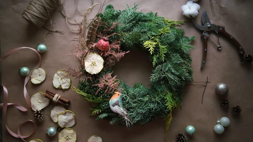 A holiday wreath made of greenery is surrounded by items used to make it -- ribbon, string, decorative balls and garden