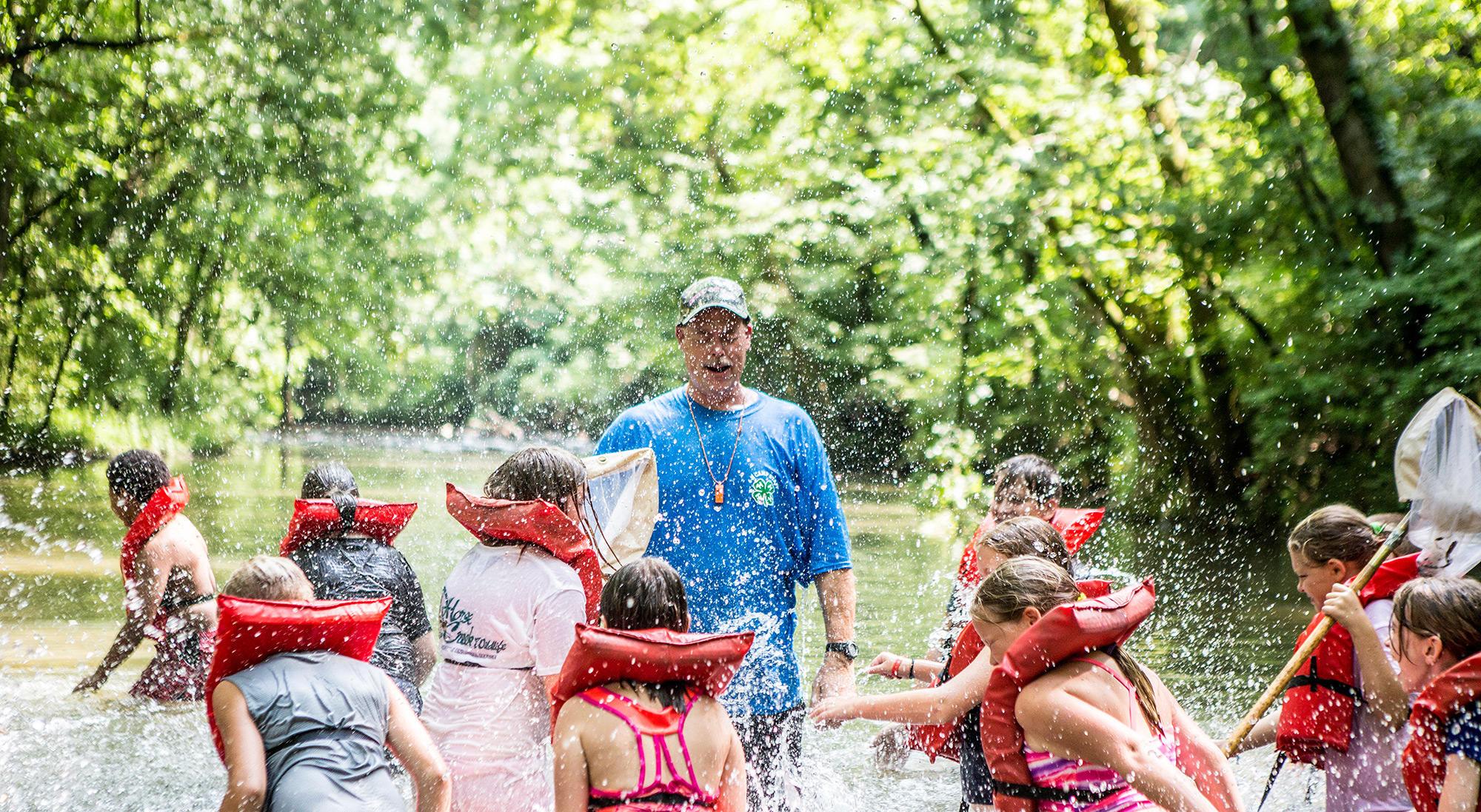 4-H members are playing and learning in the river wearing life preservers—the adult volunteer, also in the river, smiles.