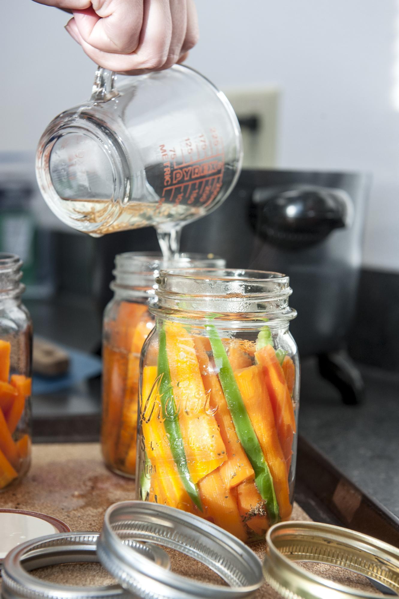 Liquid is poured from a measuring cup into a jar full of sliced carrots during a food preservation class.