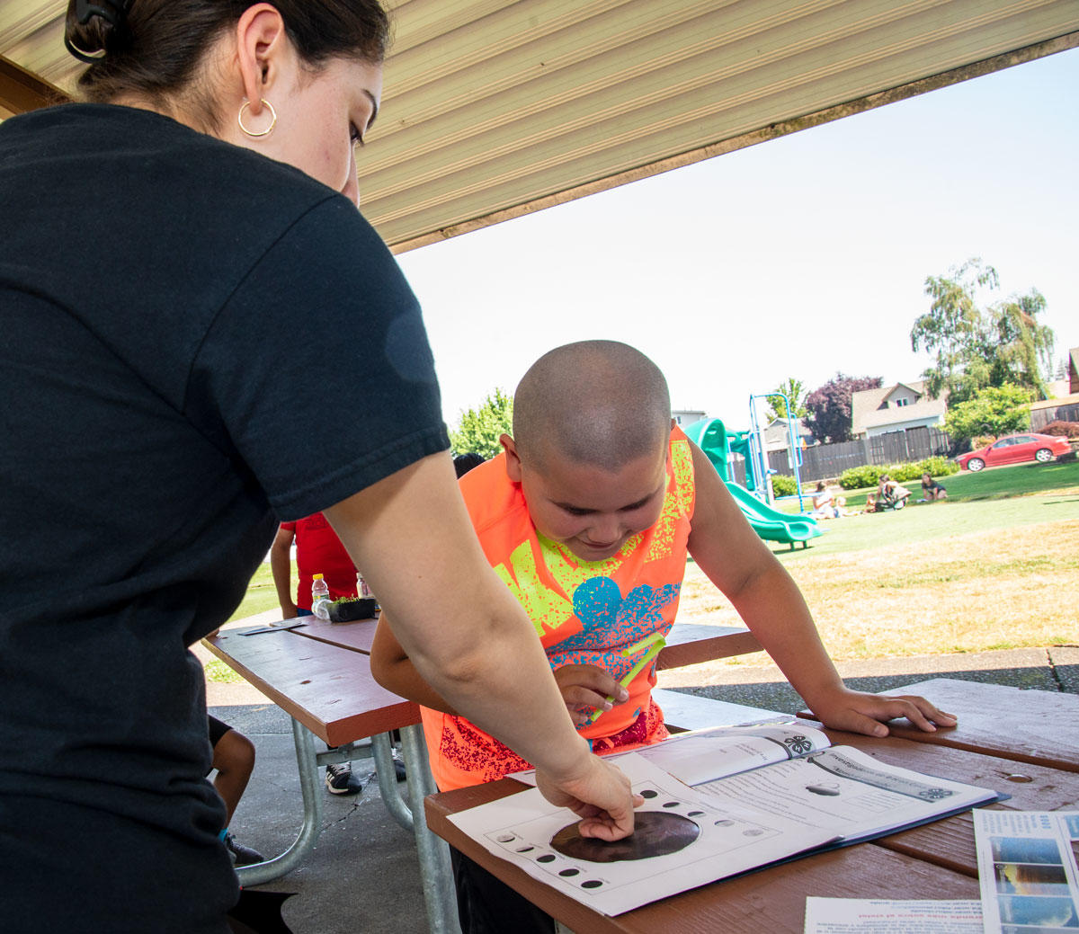 A woman in a black T-shirt points and a boy in a tie-dye T-shirt look at a piece of paper on a picinic table in a park.