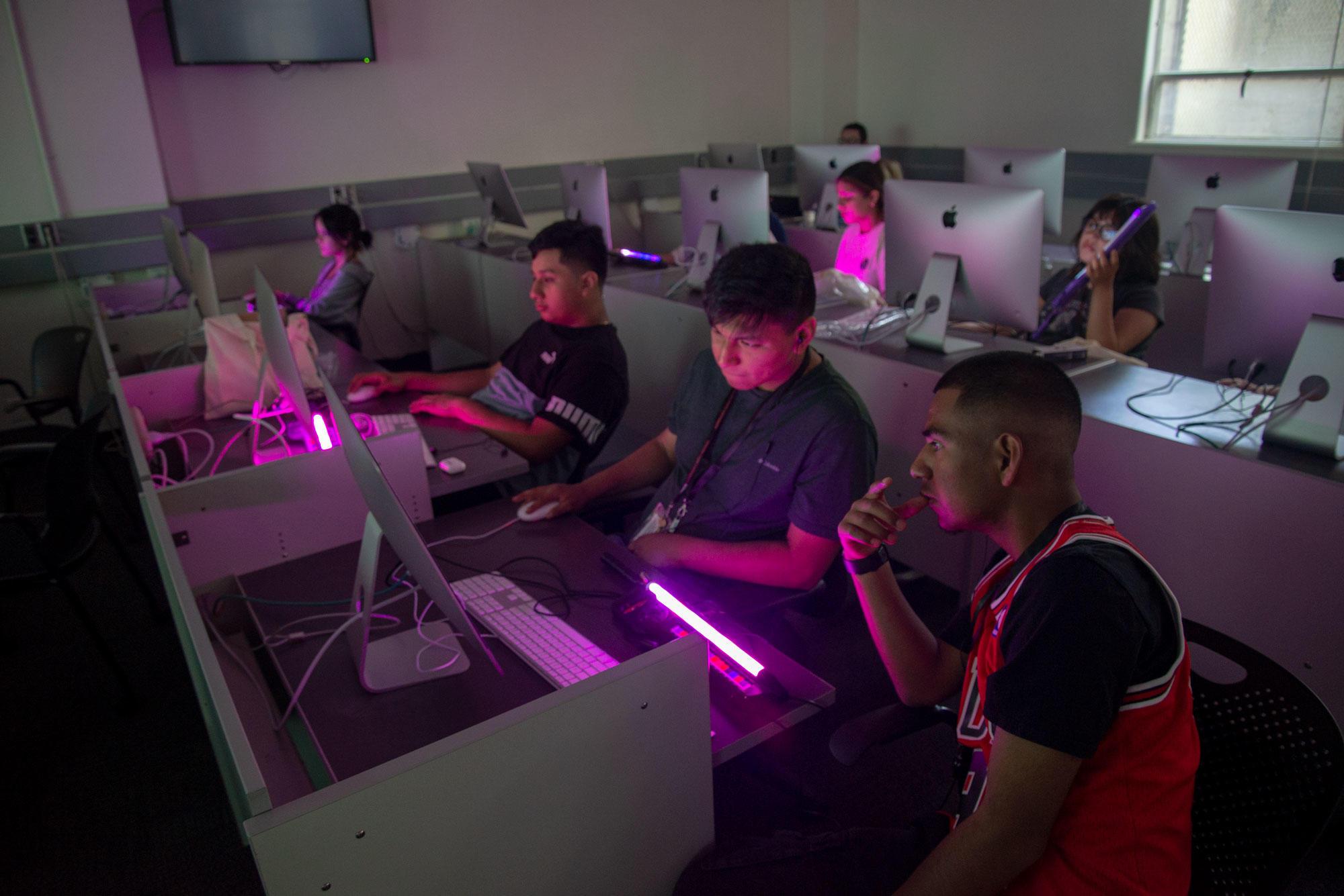 Teenagers are sitting in a computer lab for a workshop, using 3-D printed guitars that glow like light sabers.