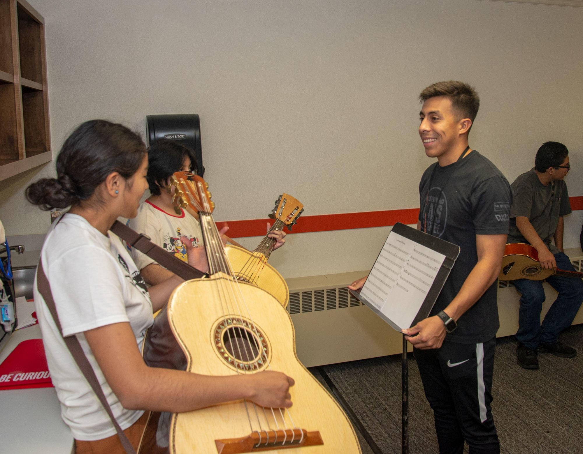 Two teenaged girls are playing large acoustic guitars while talking to a young man during a rehearsal.