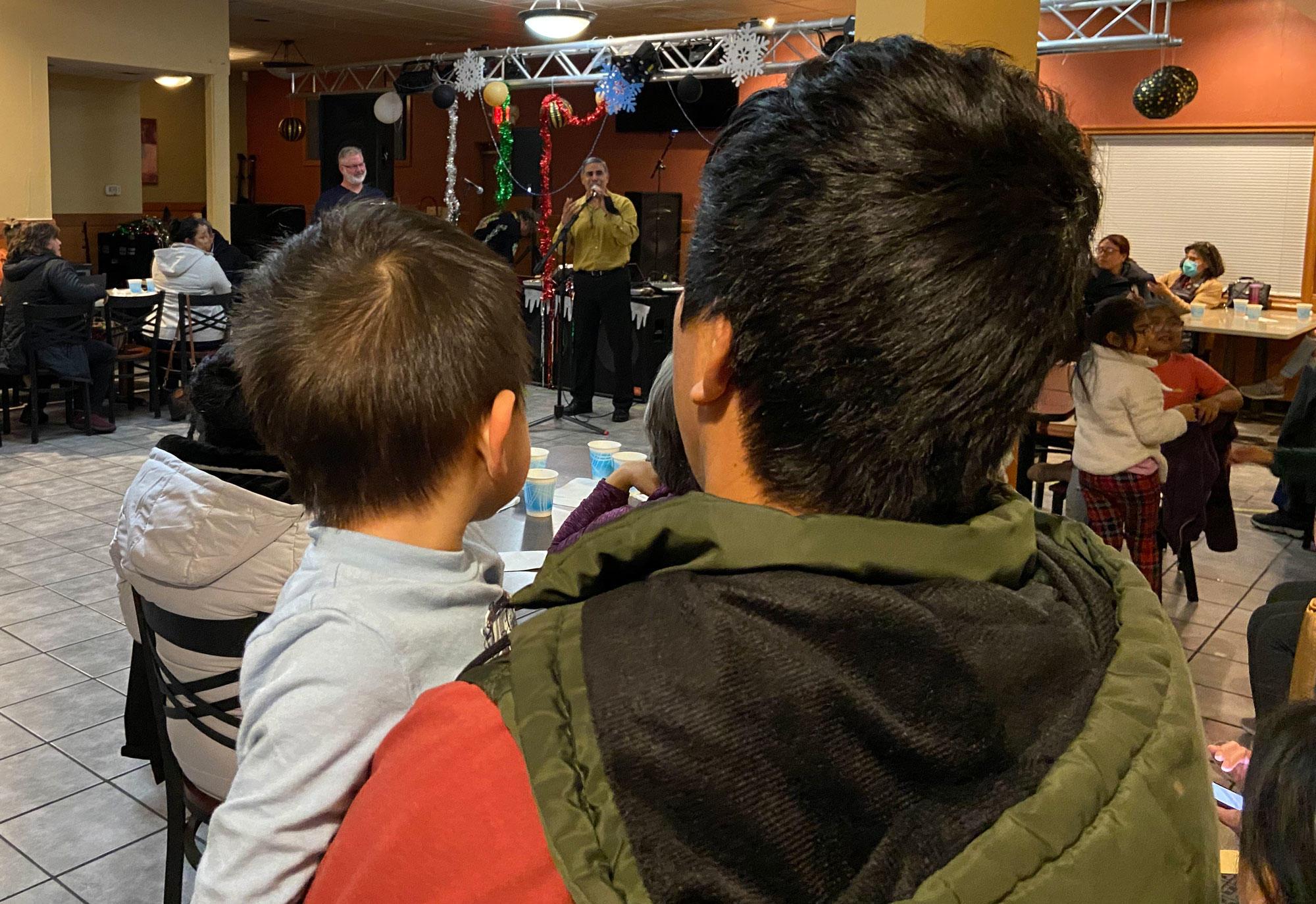 A Latino man holds a little boy while listening to a speaker at a COVID-19 town hall information event at a Mexican food restaurant.