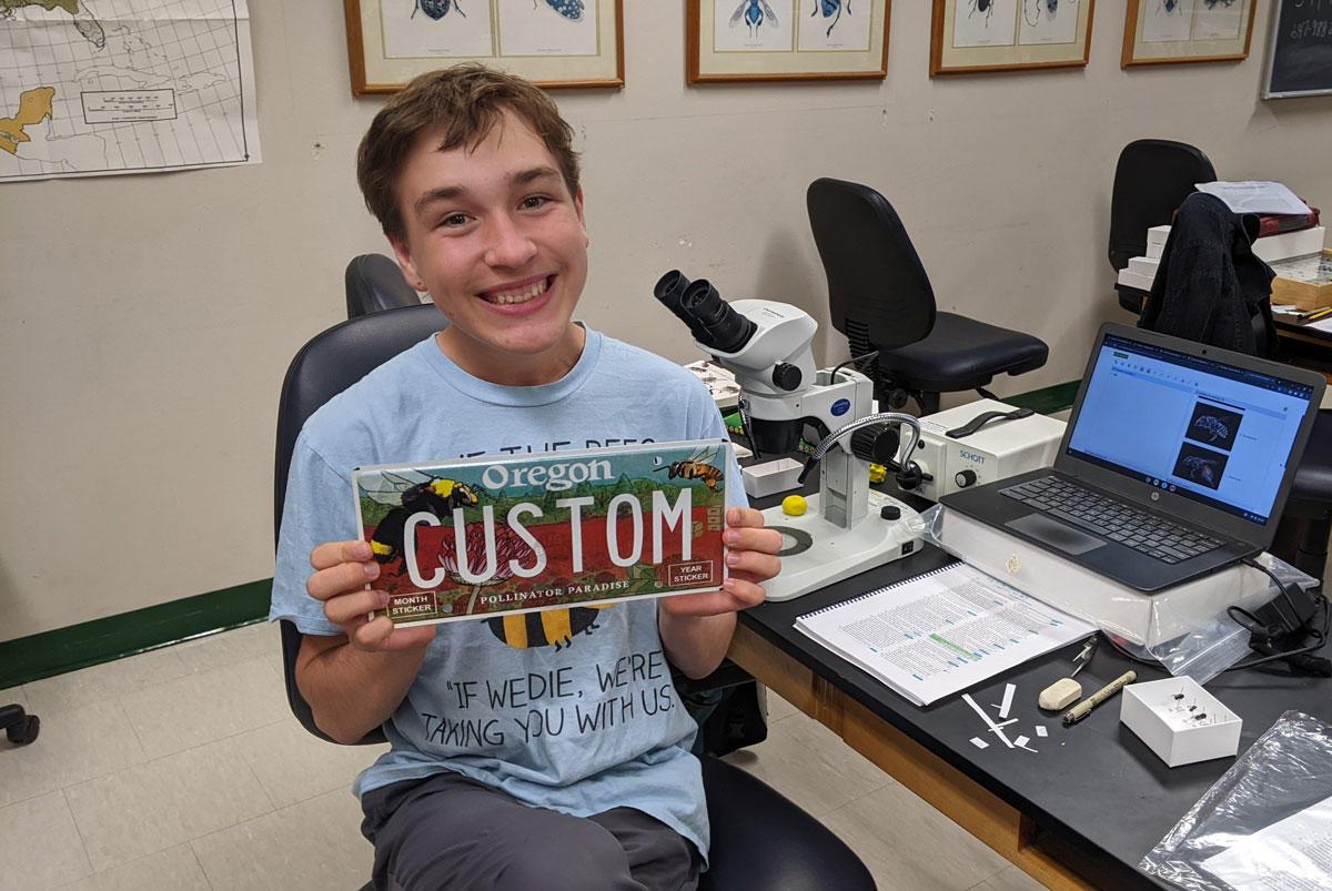 Marke Stanton sits in chair in an office and smiles as he holds up a custom pollinator license plate.