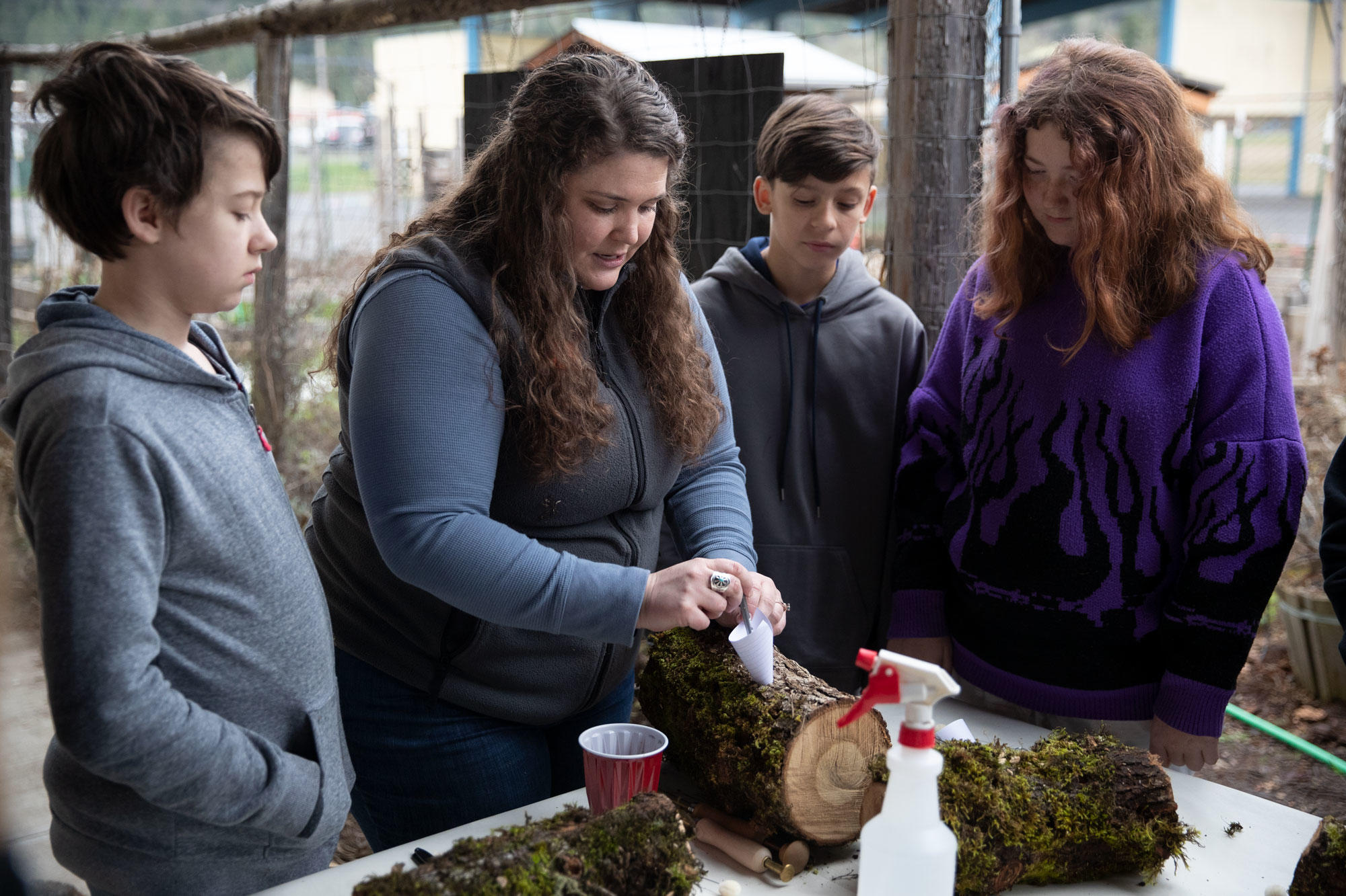 A woman uses a paper funnel to put sawdust in a hole in a log while sixth graders watch.
