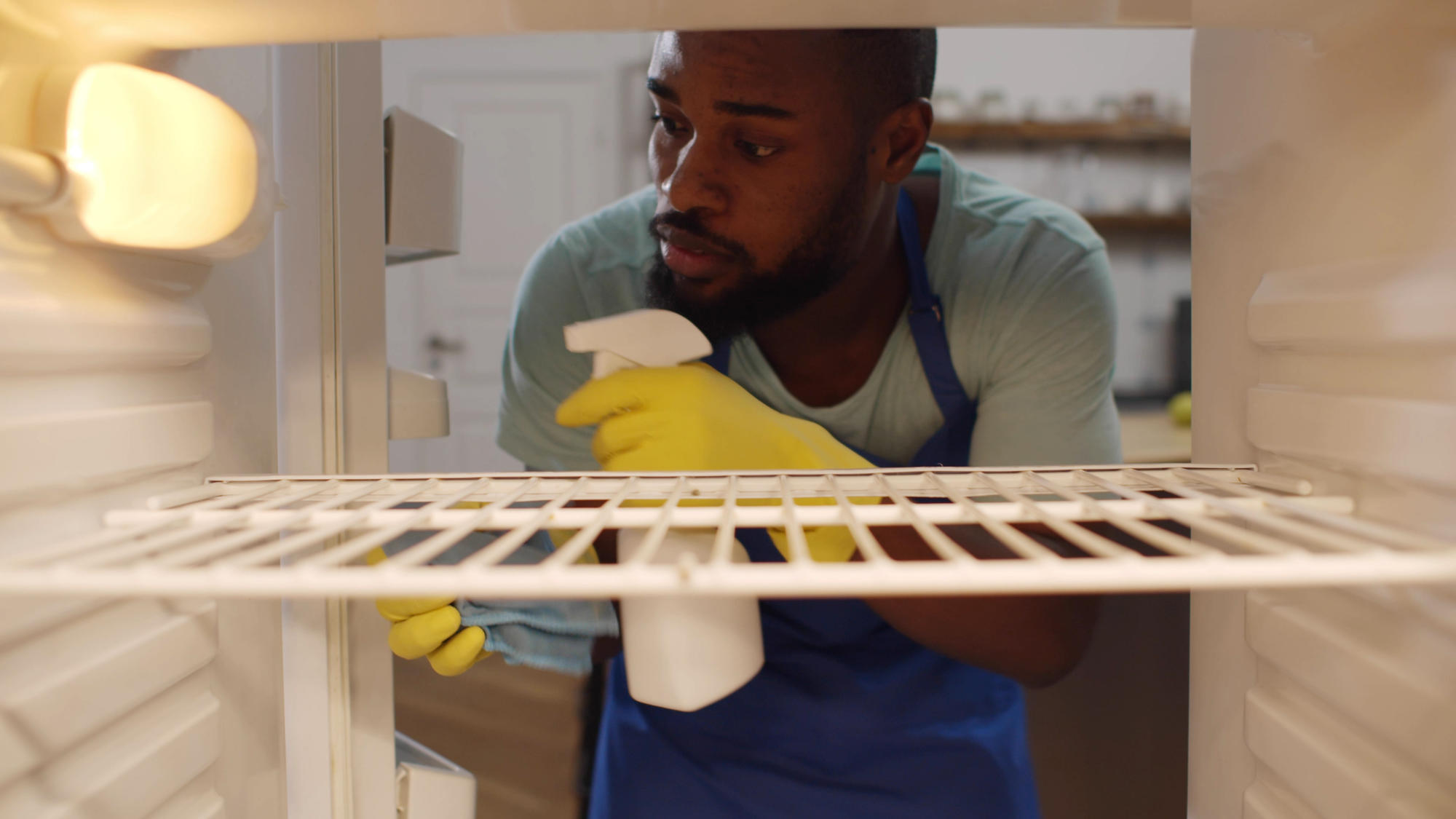 A person cleaning their fridge with a spray bottle of solution and wearing rubber gloves and an apron.