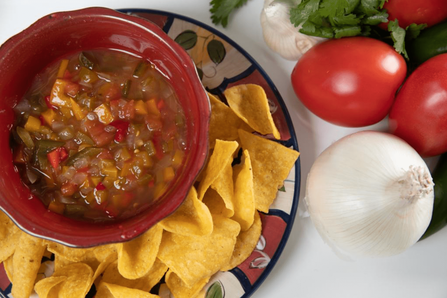 festive pottery plate with a red bowl with tomato salsa on the plate, surrounded by corn tortilla chips. White tablecloth background, next to the plate, has a white onion, roma tomatoes, cilantro, and a head of garlic partially visible from under the cilantro