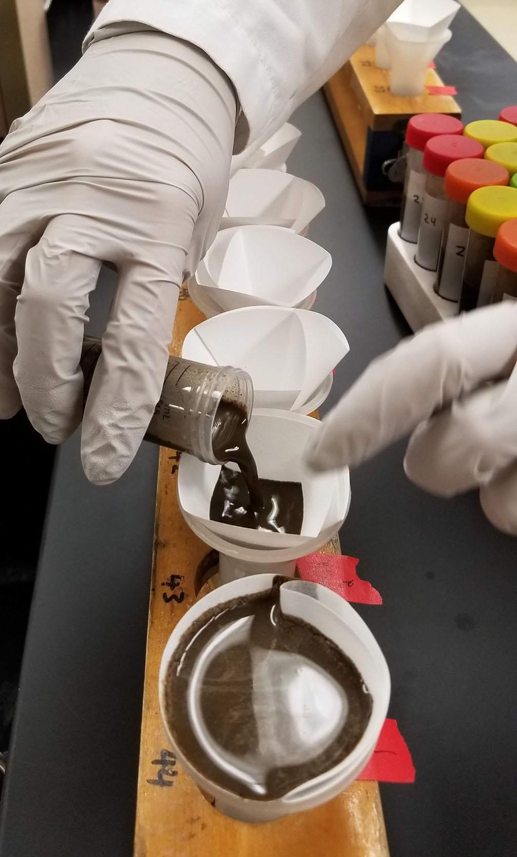 A technician in white lab coat and gloves pours black substance into row of white cups