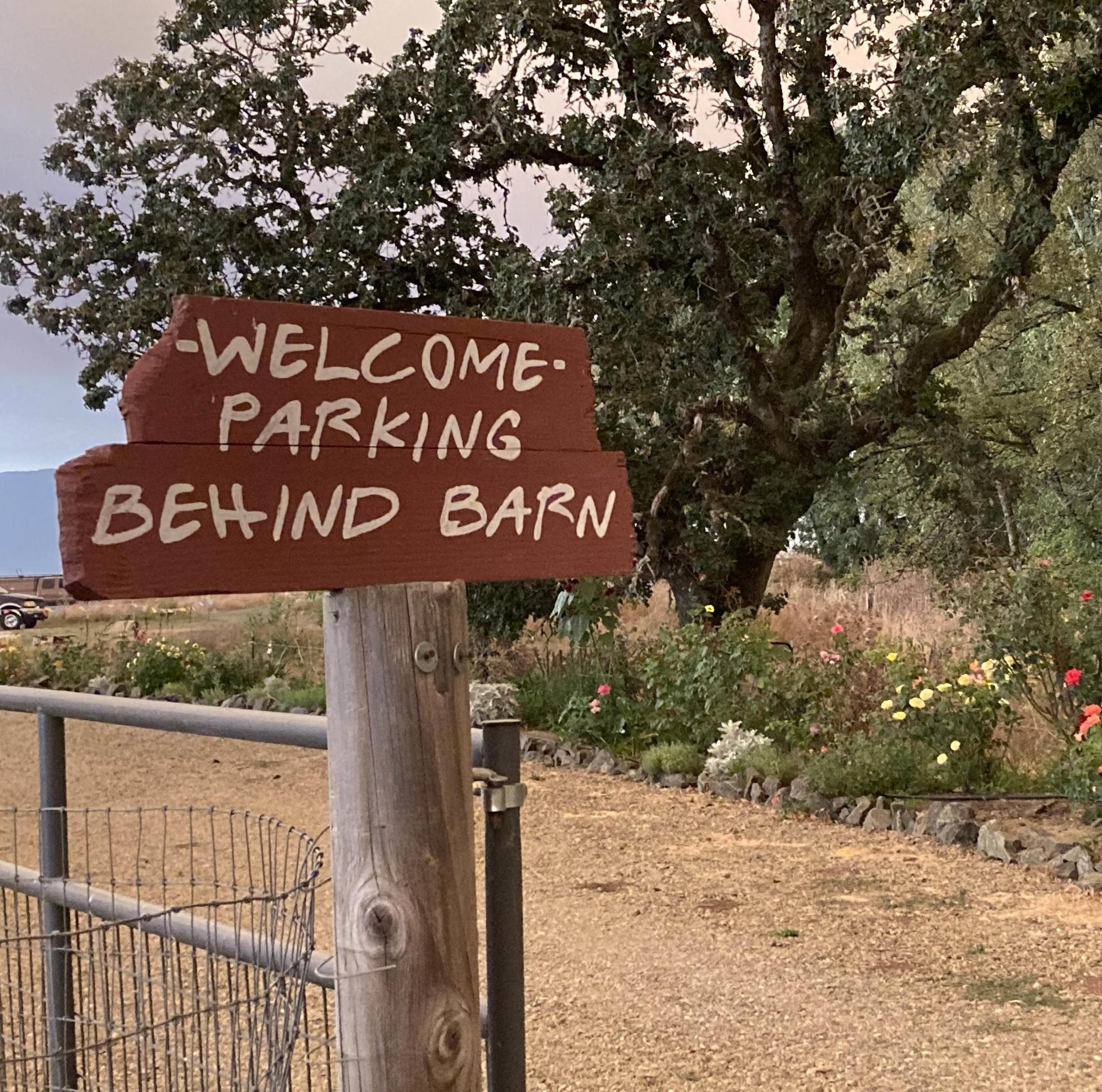 Sign reading Welcome — Parking Behind Barn on top of a post next to a dirt road. Tree and flowers in background.