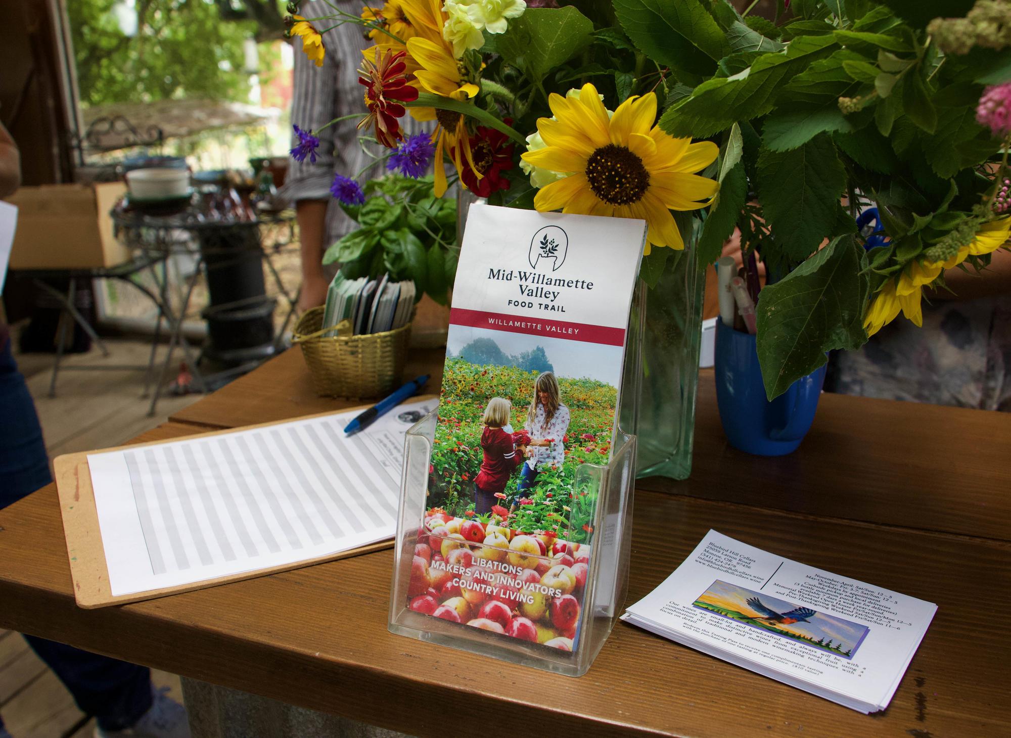 A food trail brochure, clipboard and flyer on a table in front of a bouquet of sunflowers