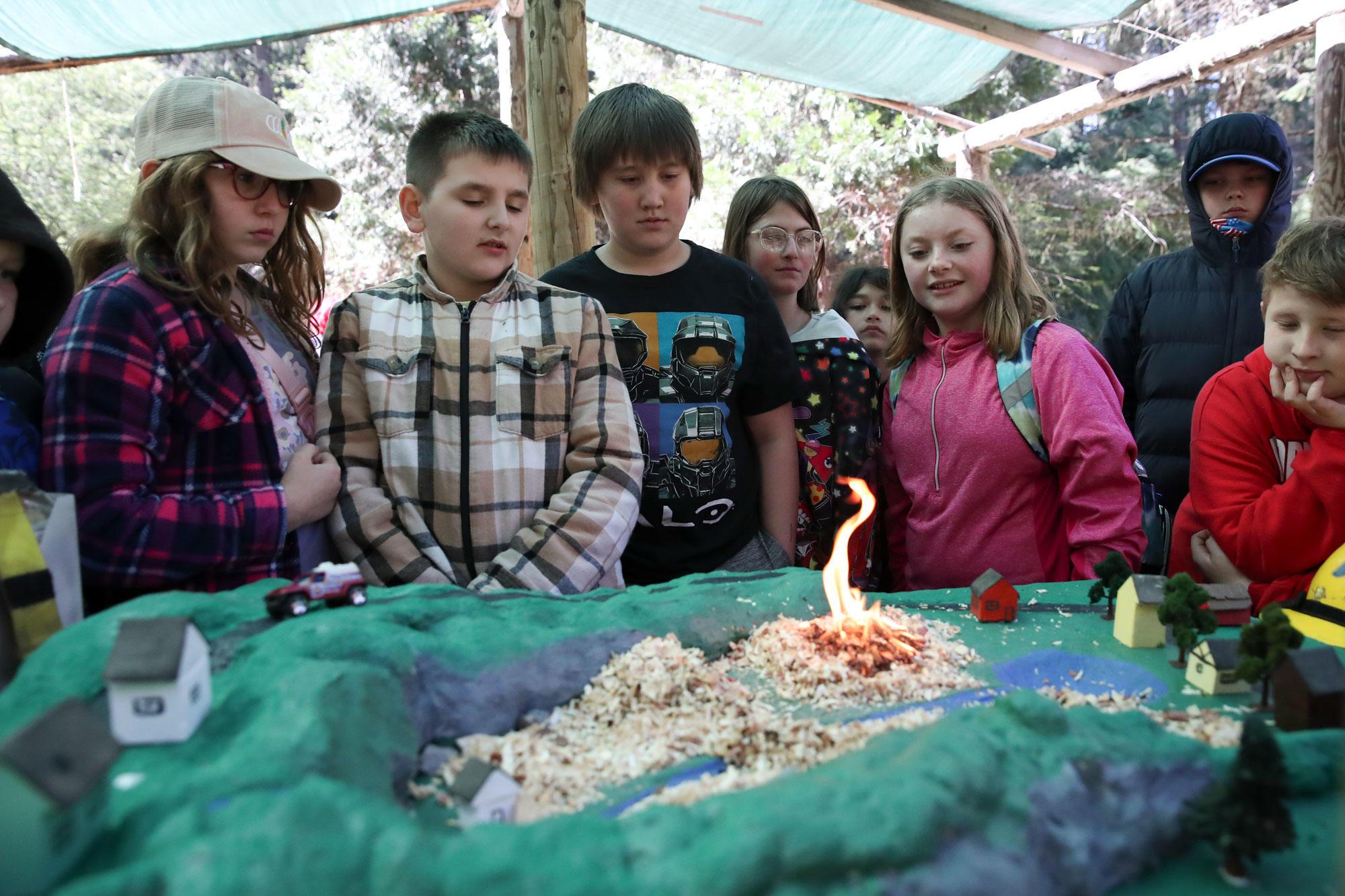 Boys and girls stand around a model of a landscape and watch a flame.
