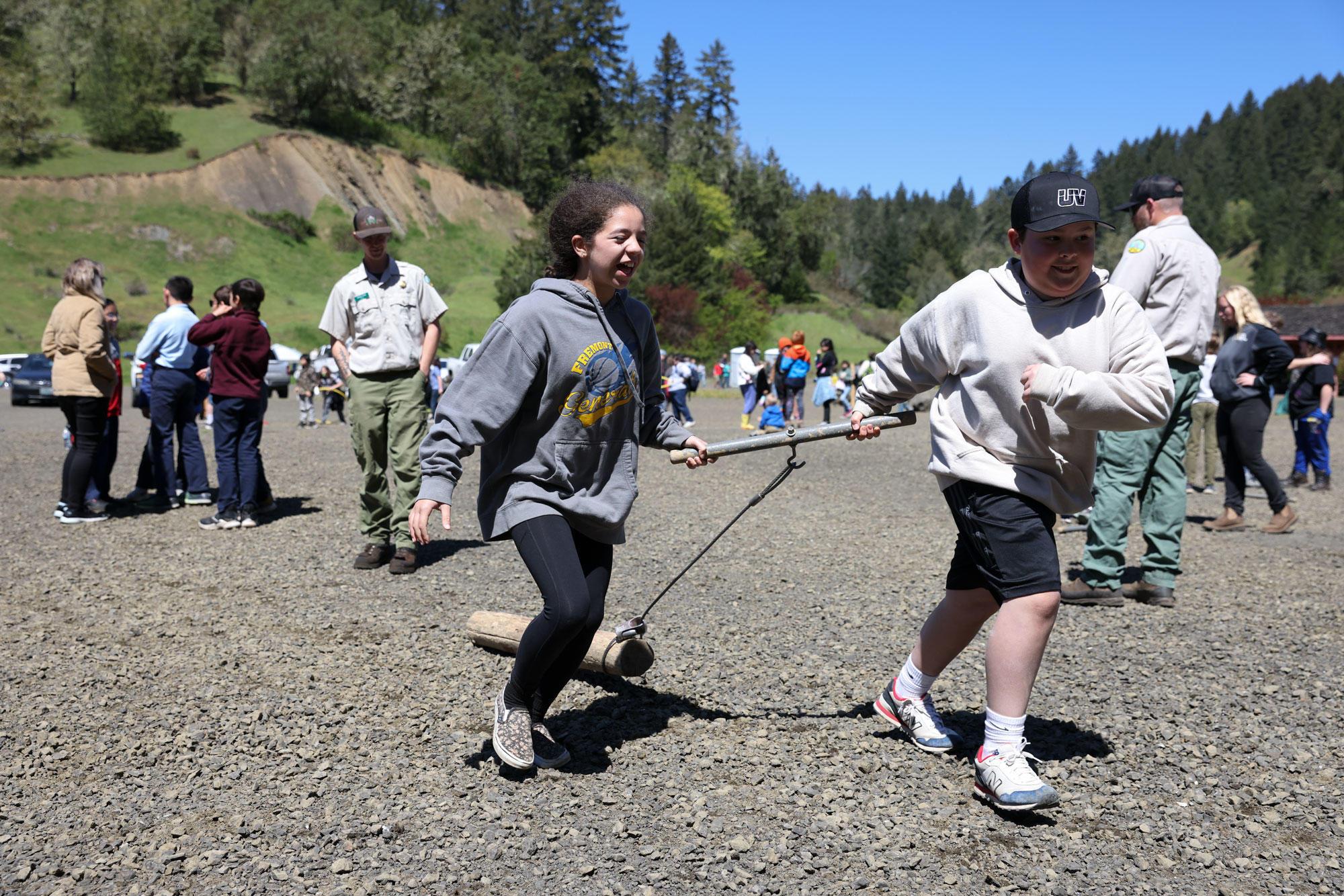 A girl and a boy use a choker cable to drag a small log across a paved parking lot.