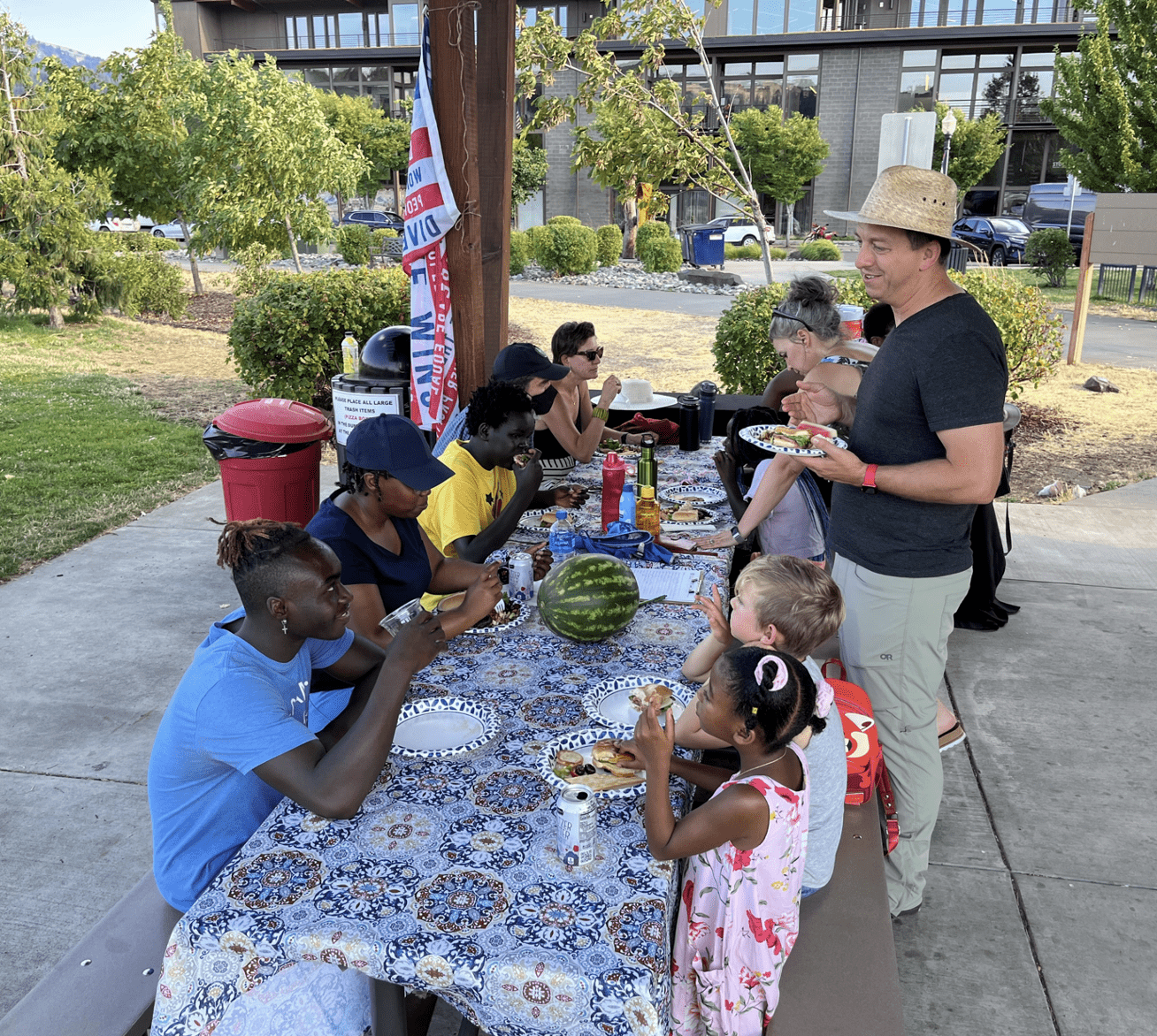 A group of people of many races sit together around a picnic table with a floral table cloth. The group eats sandwiches and many are smiling.