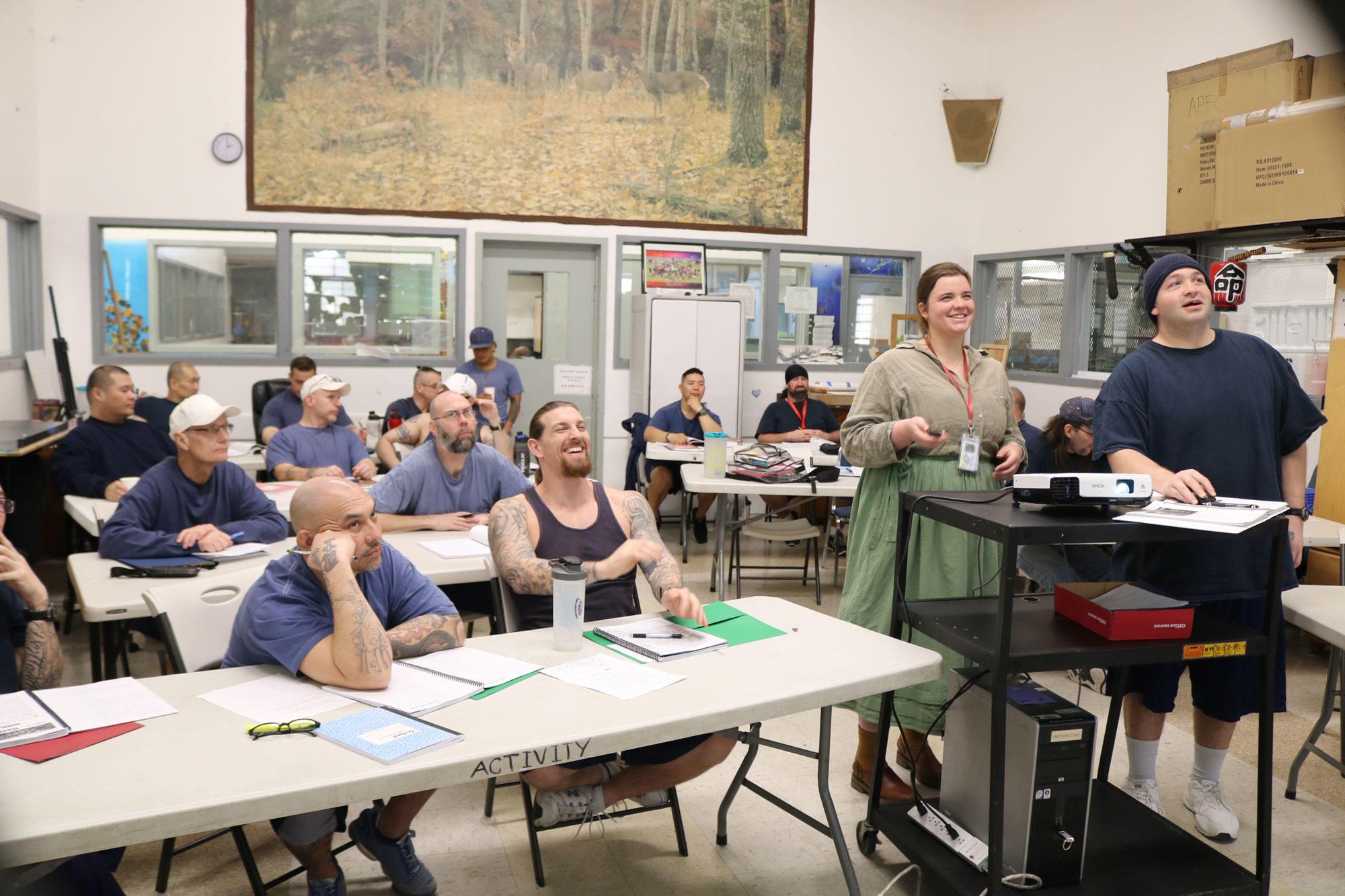 Incarcerated students sitting at tables in a classroom in the Pregon State Penitentiary are smiling as they look at a screen.