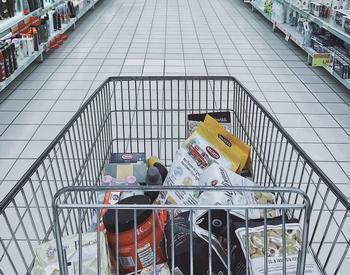 A partially filled shopping cart is depicted in the middle of an aisle between shelves in a store.