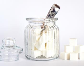 A glass jar containing sugar cubes with tongs extending out of the top of the jar. Outside the jar are its lid and six sugar cubes stacked in a pyramid shape.