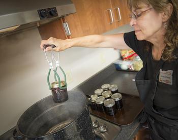 A woman stands next to a stove and removes a jar of jelly from a boiling water bath canner using a canning lifter.