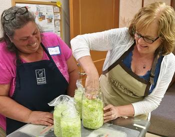 A woman packs cabbage into a jar to make sauerkraut as another woman looks on.