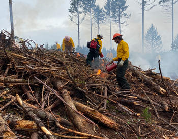 Yellow-shirted instructors from OSU Extension’s Forestry and Natural Resources Extension Program stand on a wood pile and apply accelerant to start the controlled burn.
