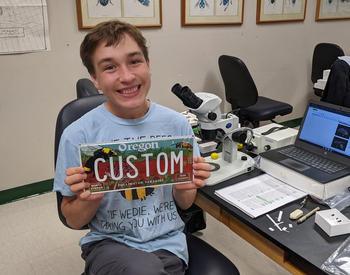 Marke Stanton sits in chair in an office and smiles as he holds up a custom pollinator license plate.