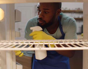 A person cleaning their fridge with a spray bottle of solution and wearing rubber gloves and an apron.