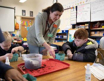 A woman stands over a table in a second-grade classroom, helping the students plant seeds in a container.