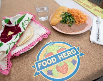 A plate of Mexican food and a plate of flour tortillas flank a Food Hero logo in a table.