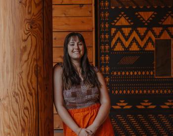 A person wearing an orange skirt and brown woven top stands next to a wooden pillar. The person has long brown hair and is smiling.