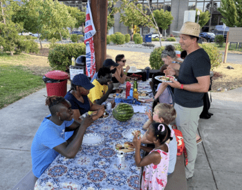 A group of people of many races sit together around a picnic table with a floral table cloth. The group eats sandwiches and many are smiling.