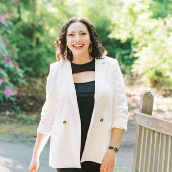 Nadine Menashe is standing outside smiling in a black dress and white blazer