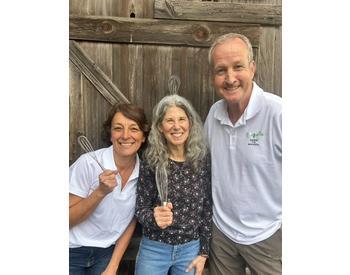 three smiling people holding whisks standing in front of a wooden fence